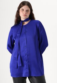 Blå satengbluse med høy krage, lange ermer og knappelukking foran. Har en bundet sløyfe ved halsen og en jevn tekstur.