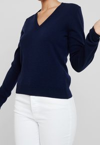 Marinblå v-ringad tröja i mjukt material, med långa ärmar och en figurnära design, som matchas med vita byxor.