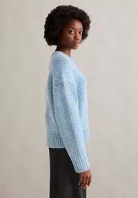 Jasnoniebieski sweter z dzianiny o luźnym kroju, z opuszczonymi ramionami, ściągaczem u dołu i teksturowanym wykończeniem. Noszony z czarną spódnicą.