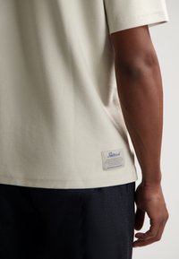 Camiseta de manga corta color beige claro con tejido texturizado y etiqueta en el dobladillo. Se lleva con pantalones cortos oscuros, mostrando un diseño sencillo.