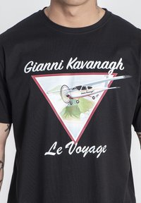 Černé bavlněné tričko s barevným grafickým motivem letadla a horské krajiny uvnitř trojúhelníkového designu. Text uvádí "Gianni Kavanagh" a "Le Voyage."