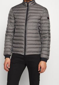 Grå pufferjakke med horisontalt quiltet mønster, høj krave, frontlynlås og elastiske manchetter. Ingen synlig branding eller mønstre.