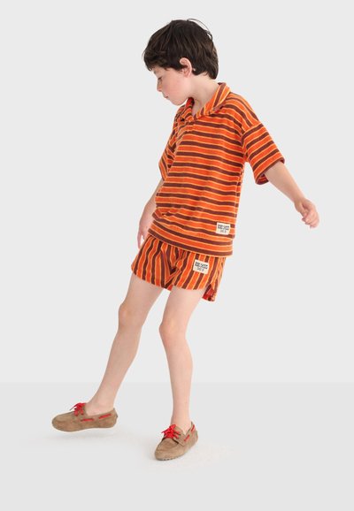 Garçon en équilibre sur un pied, portant un t-shirt et un short rayés orange et marron, avec des mocassins beige à lacets rouges.