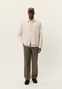 Ανοιχτό μπεζ βαμβακερό μπουφάν με κουμπιά στιγμής, δύο μπροστινές τσέπες, συνδυασμένο με καφέ υφασμάτινο παντελόνι με υφή και καφέ καπέλο. Τα καφέ παπούτσια ολοκληρώνουν την εμφάνιση.