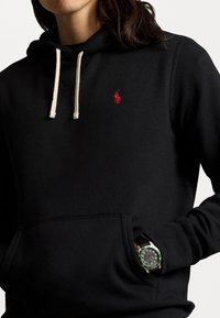 Svart hoodie med en liten röd logotyp, med beige snoddar och en framficka. En grön klocka med silverarmband är delvis synlig.