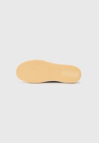 Semelle de chaussure en caoutchouc beige avec un motif discret en zigzag sur un fond gris clair uni.