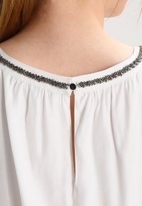 Blouse blanche avec un col rond agrémenté de perles métalliques et d'une fermeture à bouton à l'arrière, avec des détails en tissu froncé.