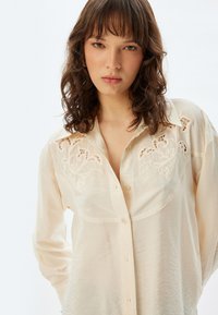 Ivory blouse met een knoopsluiting aan de voorkant, geborduurde bloemdetails bij de schouders en een gestructureerde stof. Lange mouwen, comfortabele pasvorm.