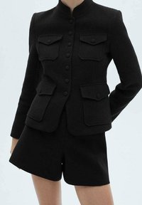 Veste noire texturée avec poches à rabat boutonnées, portée avec un short noir assorti, présentée sur une personne devant un fond uni.