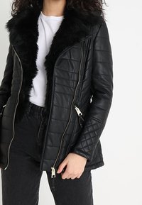 Chaqueta negra de cuero acolchada con cuello de piel sintética, con cremalleras plateadas y una forma ligeramente entallada. Se lleva sobre una camiseta blanca.