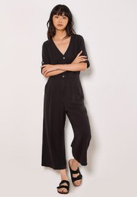 Mono negro con un escote en V profundo, mangas cortas, botones en la parte delantera y piernas anchas hasta los tobillos. Combinado con sandalias negras. Textura de tejido suave.