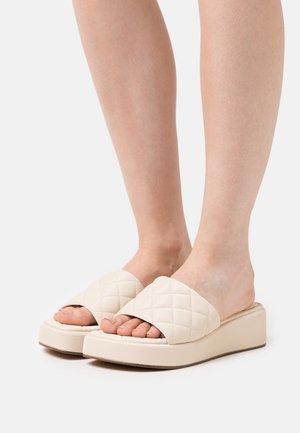 Pies que llevan sandalias plataforma de color crema acolchonadas, abiertas en la punta, con suelas gruesas sobre un fondo blanco.