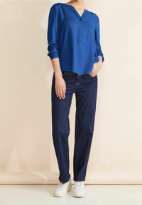Blusa blu a maniche lunghe con scollatura a V e tre bottoni, abbinata a jeans in denim scuro e sneakers bianche. Tessuto dalla superficie liscia.