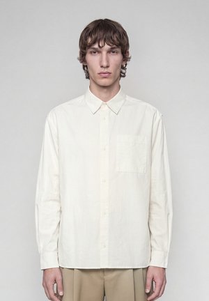 Junger Mann mit mittellangen braunen Haaren, der ein schlichtes weißes Hemd mit Knöpfen und beige Hose trägt, steht vor einem grauen Hintergrund.