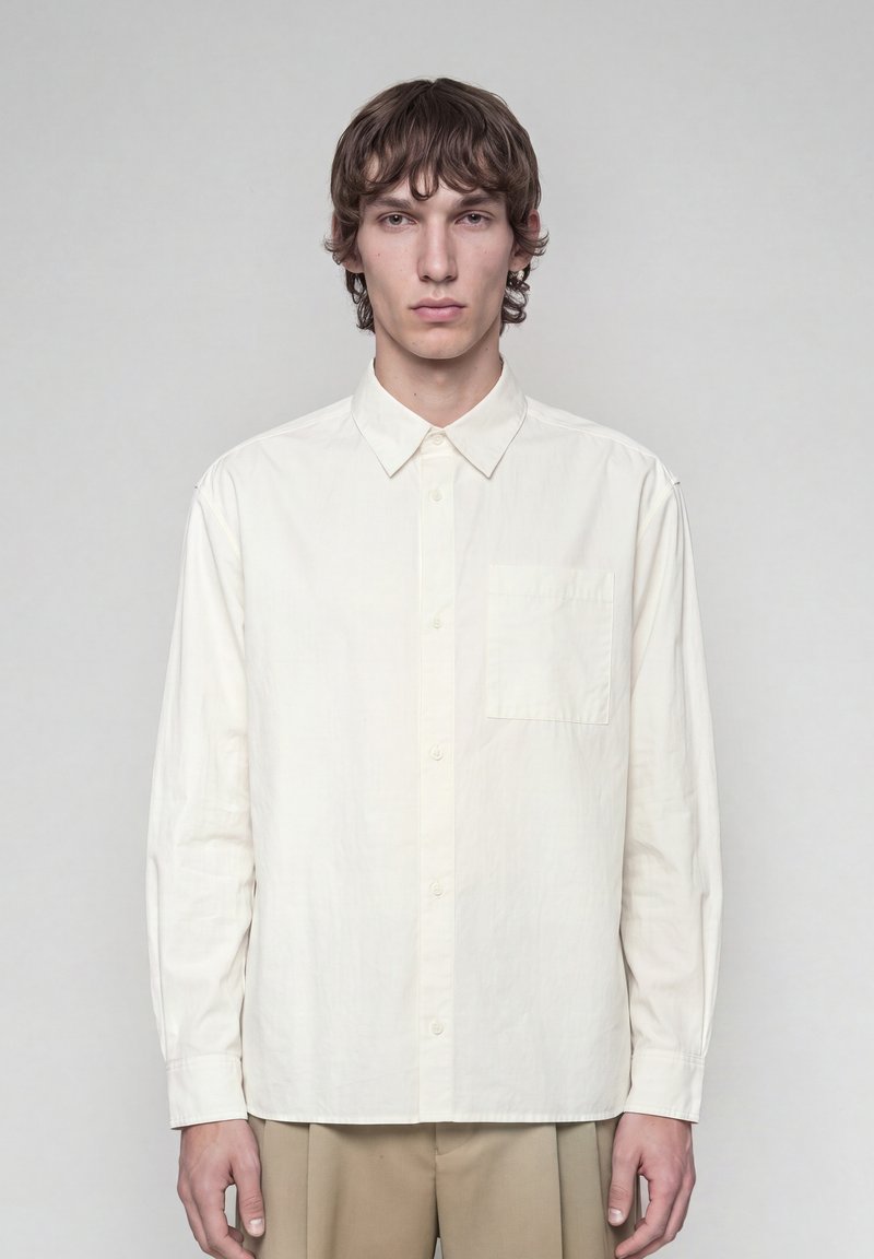 Jeune homme aux cheveux bruns mi-longs, portant une chemise blanche unie boutonnée et un pantalon beige, debout devant un fond gris.