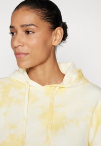 Hoodie jaune tie-dye avec cordon de serrage, présentant une texture douce, une coupe décontractée et une grande capuche. L'arrière-plan est blanc uni.