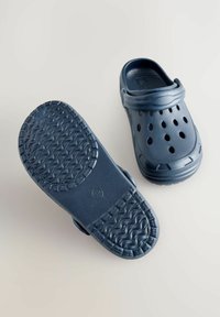 Marinblå plastträsko med strukturerad sula, ventilationshål och en justerbar rem. Har en rund tå och en slät yta.