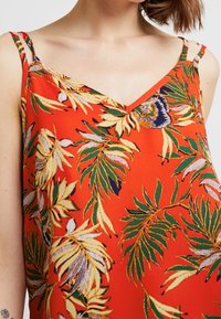 Röd ärmlös topp med V-ringning, prydd med ett tropiskt bladmönster i grönt och gult. Tunn axelremmar och lätt material.