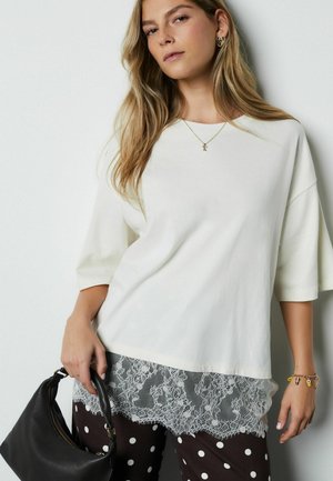 Biela nadrozmerná tričko s krátkymi rukávmi, cez ktoré je vrstva so sivým čipkovaným lemom. Kombinované s hnedými nohavicami s bodkami a čiernou kabelkou.