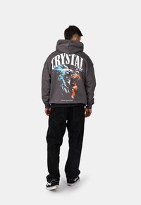 Felpa grigia con vestibilità oversize caratterizzata da una grafica di un leone colorato e testo "CRYSTAL PARIS" sulla schiena; abbinata a pantaloni neri e sneakers.