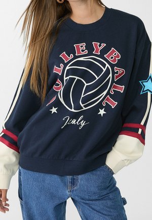 Vrouw die een marineblauwe sweatshirt draagt met volleybalafbeelding, tekst "VOLLEYBALL Italy", crèmewitte mouwen met rode strepen, blauwe ster patch en blauwe spijkerbroek.