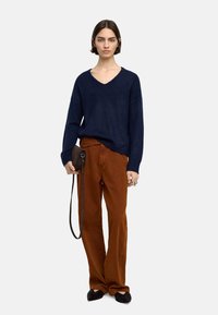 Niebieski sweter w serek, luźny krój, z rudymi spodniami o prostym kroju. Modelka trzyma ciemnobrązową torbę na ramię z paskiem.