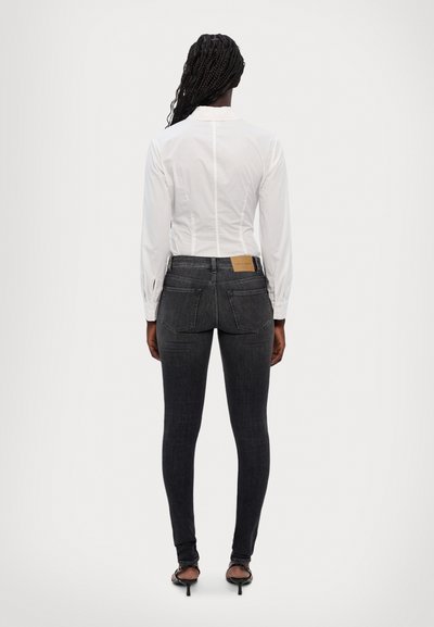 Jeans skinny de culoare gri închis, cu cinci buzunare, talie înaltă și decolorare subtilă. Asortate cu o cămașă albă cu mânecă lungă, prezentate din spate.