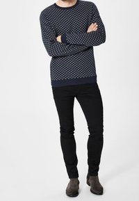 Man som står med armarna i kors, iförd en marinblå mönstrad tröja, svarta slim-fit byxor och bruna mockakängor mot en plain bakgrund.