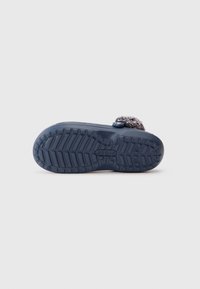 Mørkeblå Crocs slippers med struktureret sål og fluffy, grålig overdel synlig fra underliggende perspektiv mod en hvid baggrund.