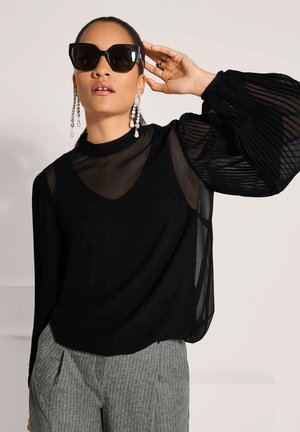 Vrouw met oversized zwarte zonnebril, lange hangende parel oorbellen, doorschijnende zwarte blouse met pofmouwen en grijze pantalon met krijtstreep.