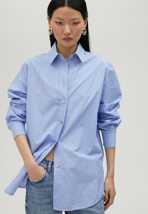 Camicia button-up a righe blu chiaro con un fit rilassato, caratterizzata da dettagli decorativi in argento e maniche arrotolate. Abbinata a jeans in denim.