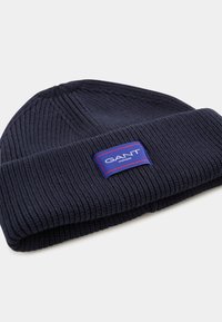 Dark blue knitted beanie made of ribbed fabric with a folded cuff. Features a rectangular GANT logo patch in red and white on the front.
