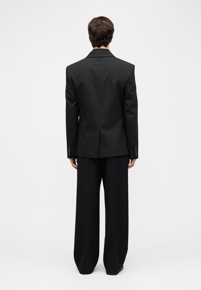 Personne portant un blazer noir et un pantalon noir à jambes larges, debout de dos sur un fond blanc uni.