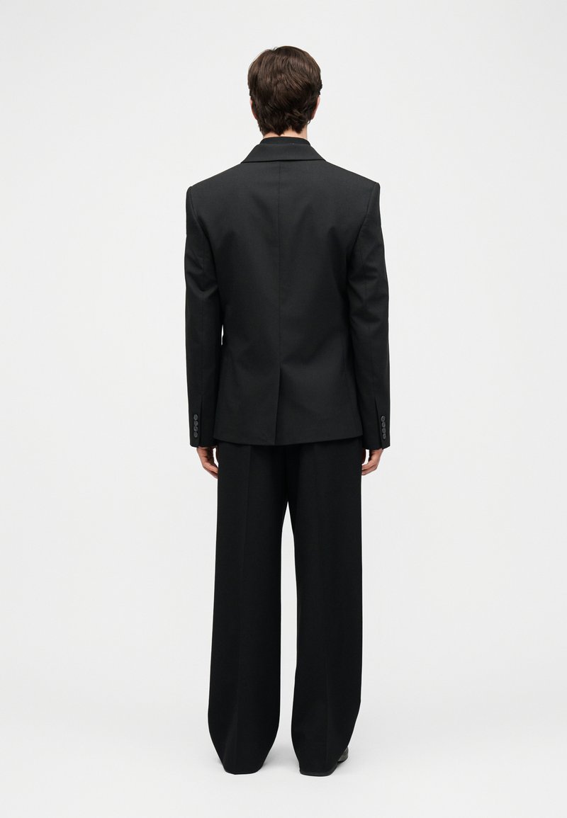 Person iført en sort blazer og sorte bukser med vide ben, stående med ryggen vendt mod en ensfarvet hvid baggrund.