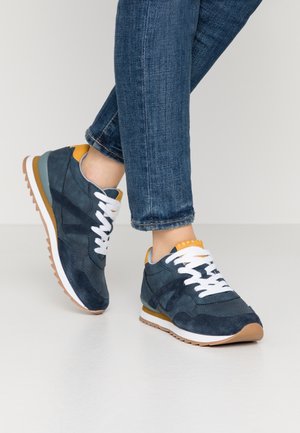 Laivastonsiniset ja sinappikeltaiset tennarit, joissa on valkoiset kengännauhat, puettuna ylös käärittyjen sinisten farkkujen kanssa, esitettynä vaalealla, sileällä lattiapinnalla.