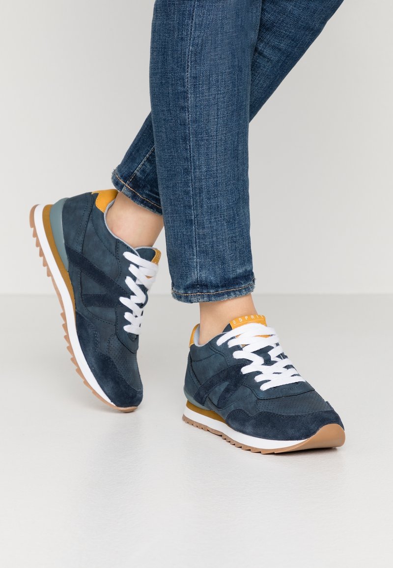 Baskets bleu marine et moutarde avec des lacets blancs portées avec un jean bleu retroussé, posées sur une surface de sol claire et lisse.