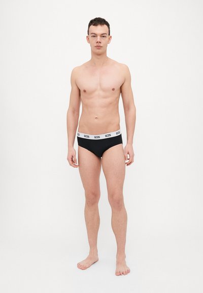 Young man standing barefoot wearing black briefs with a white waistband labeled GCDS against a plain white background.