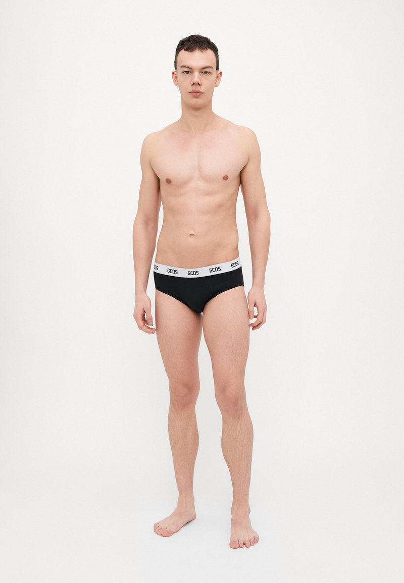 Black underwear with white "GCODS" logo on the elastic waistband. Model stands barefoot and poses frontally against a plain white background.