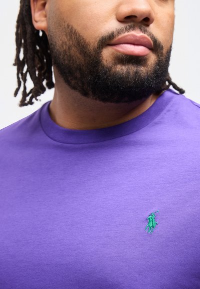 Άνδρας με γενειάδα και ντραντλoκς, φορώντας μοβ μπλουζάκι με στρογγυλή λαιμόκοψη κ αι μικρό πράσινο λογότυπο παίκτη πόλο στο στήθος, κοιτάζει προς τα δεξιά.