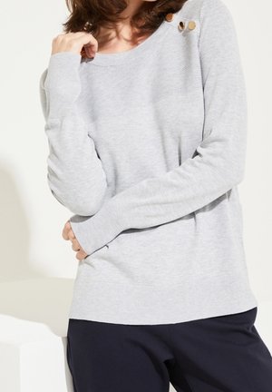 Suéter de punto gris claro con mangas largas, que presenta un escote redondo y tres botones decorativos en el hombro. Textura suave, corte casual.