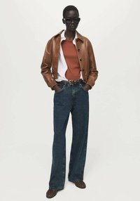 Rjava usnjena jakna preko vrhnjega dela v barvi rje, kombinirana z ohlapnimi modrimi kavbojkami in rjavimi čevlji. Očala za sonce dopolnjujejo opravo.