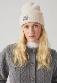 Beanie beige lavorato a maglia con texture a coste e un'etichetta. Cardigan grigio a trecce con bottoni tondi e un dolcevita bianco sotto.