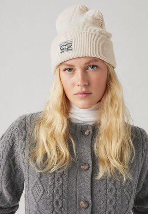 Beige gebreide beanie met een geribbelde textuur en een label. Grijze kabelgebreide cardigan met ronde knopen en een witte coltrui daaronder.
