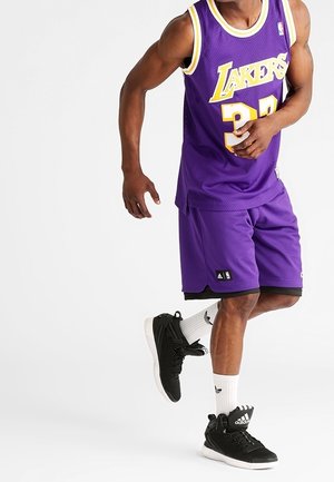 Man wearing purple Lakers basketball jersey and shorts, white Adidas socks, and black Adidas sneakers, standing with one foot raised.