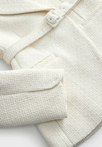 Veste tissée texturée couleur crème avec une ceinture assortie et une fermeture à un seul bouton, posée à plat sur un fond clair.