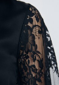 Abito nero con un corpino in satin liscia e una manica in pizzo trasparente con intricati motivi floreali e bordi scallopati.