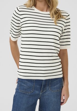 Vrouw die een wit kortärmelig truitje met zwarte horizontale strepen en blauwe spijkerbroek draagt, staand tegen een effen achtergrond.