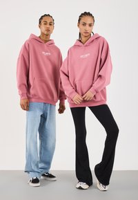 To personer med flettehår iført store rosa "OFF LIMITS" hettegensere; en i lyseblå jeans og svarte sneakers, den andre i svarte bukser og hvite sneakers.