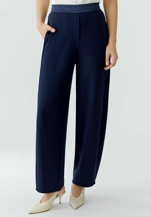 Femme portant un pantalon large bleu marine à taille élastique et des escarpins blancs à bouts pointus avec bride arrière, debout, une main dans la poche.