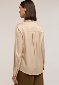 Beżowa satynowa bluzka o luźnym kroju, z dekoracyjnym kołnierzykiem zdobionym ćwiekami oraz mankietami. Z tyłu znajduje się plisowanie, które zwiększa swobodę ruchów.